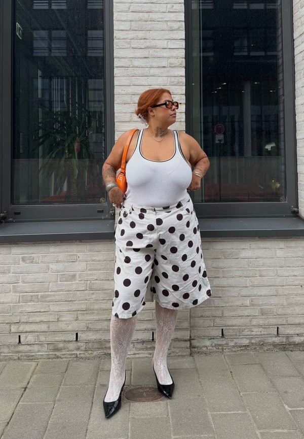 Frau mit roten Haaren, die Sonnenbrille, ein weißes Tanktop, gepunktete Shorts, Spitzentights und schwarze Absätze trägt, steht an einer weißen Ziegelmauer mit Fenstern.