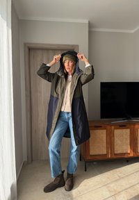 Femme à l'intérieur ajustant la capuche d'un long manteau vert foncé et bleu marine, portant un bonnet, un pull léger, un jean bleu et des bottes marron près d'une télévision et d'un meuble en bois.