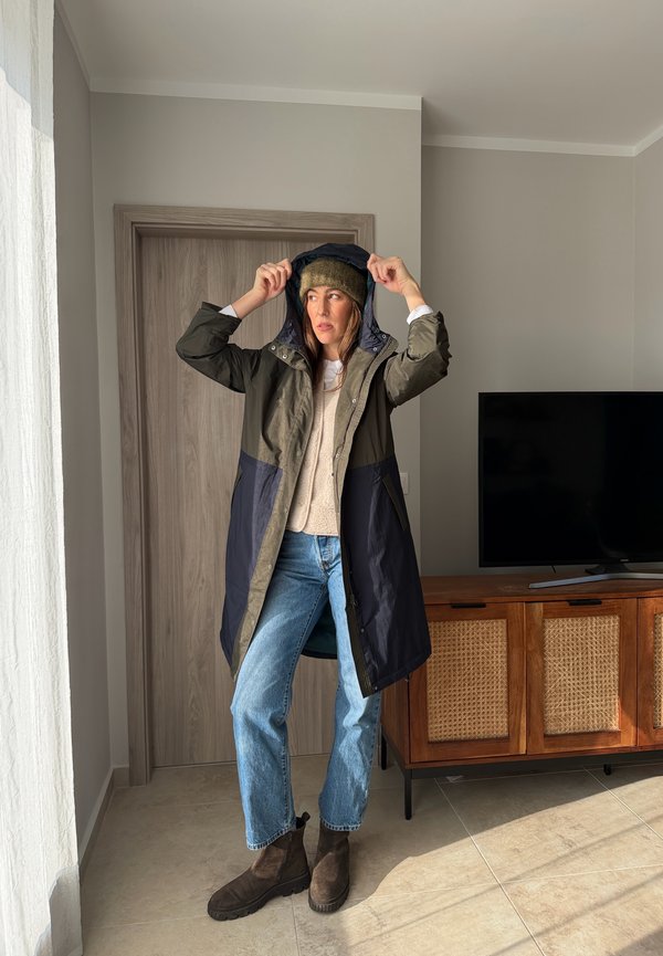 Femeie în interior care își ajustează gluga la o haină lungă verde închis și bleumarin, purtând căciulă, pulover deschis la culoare, blugi albaștri și cizme maro, lângă televizor și o comodă din lemn.