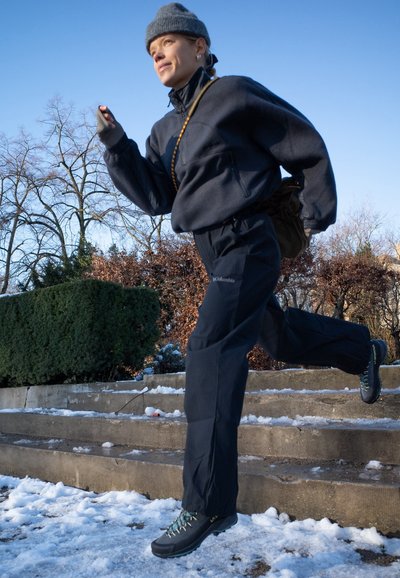 Pantalones exteriores negros y un pulóver a juego, combinados con botas negras resistentes con acentos turquesa, sobre escalones cubiertos de nieve.