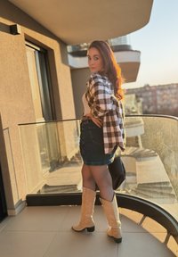 Hnědá a bílá kostkovaná flanelová košile, bílý top, tmavá džínová sukně, světle hnědé vysoké kozačky po kolena a černá kabelka na balkóně při západu slunce.