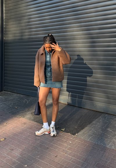 Mujer con el pelo trenzado ajusta su rostro mientras está de pie, lleva una chaqueta marrón, falda vaquera, calcetines blancos, zapatillas y sostiene una bolsa negra junto a una puerta con persiana.