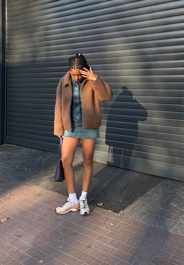 Žena s pletenými vlasmi si upravuje tvár, stojí, má na sebe hnedú bundu, rifľovú sukňu, biele ponožky, tenisky a drží čiernu tašku pri roletej dverí.