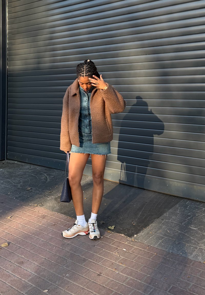 Woman with braided hair adjusts face while standing, wearing brown jacket, denim skirt, white socks, sneakers, and holding black bag by shutter door.