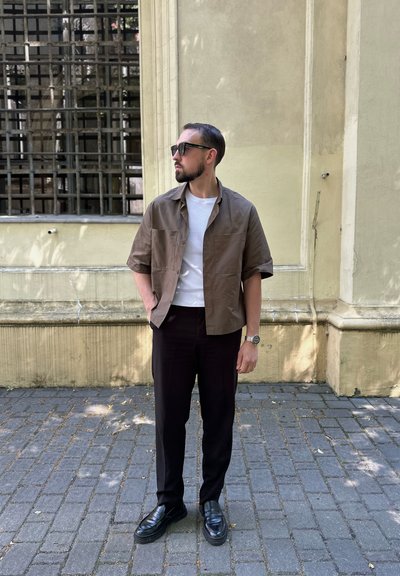 Camisa marrón de manga corta sobre una camiseta blanca, combinada con pantalones negros y zapatos negros. Al aire libre, con un fondo de pared texturizada.