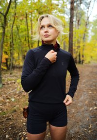 Juodas ilgomis rankovėmis, su užtrauktuku, sportinis viršus su tekstūruotu paviršiumi, derinamas su juodais prigludusiais šortais. Lauko aplinka su rudens lapija.