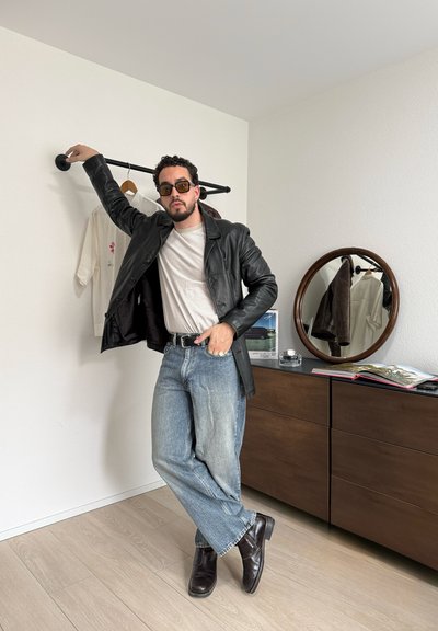 Hombre con gafas de sol, chaqueta de cuero negra, camisa a rayas, vaqueros y botas negras apoyado en una barra para ropa montada en la pared en una habitación minimalista.