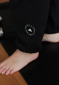 Zwarte sportbroek met een strakke boord heeft een cirkelvormig wit logo met een driehoek, in combinatie met een blote voet op een zwarte mat.