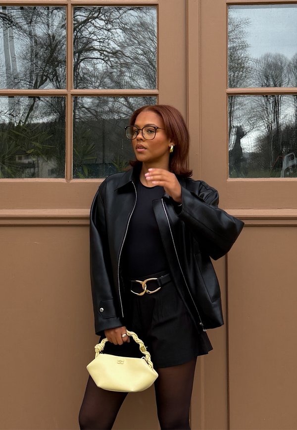 Kvinde iført sort læderjakke og briller, der holder en bleg gul håndtaske, stående foran en brun dør med glasruder.