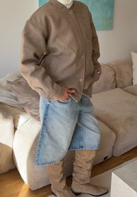 Veste aviateur en mélange de laine marron avec boutons-pression, short en denim bleu clair, et bottes beige montant jusqu'aux genoux, debout près d'un canapé.