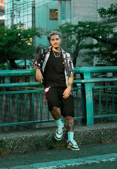 Camiseta negra, camisa de manga corta con estampado, pantalones cortos negros, zapatillas con acentos amarillos. Los accesorios incluyen collares y un llavero con funda rosa.