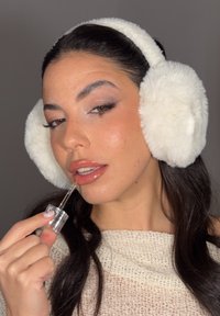 Białe nauszniki z sztucznego futra, osoba nakładająca przezroczysty błyszczyk do ust za pomocą pipety, ubrana w kremowy, teksturowany sweter, z ciemnymi włosami i subtelnym makijażem.