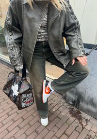Grå läderjacka med snörningsdetaljer, randig topp, mörkgröna jeans, svarta och orange Nike-skor samt en mönstrad handväska med ikoniska grafiska motiv.