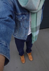 Personne portant une veste en denim bleue, un jean bleu, des bottes couleur fauve et une écharpe rayée verte et bleue debout sur un béton gris.