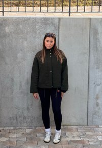 Prešita zelena jakna z visokim ovratnikom in zlatimi gumbi, kombinirana s črnimi pajkicami in belimi supergami. Lasje spusti, stoji ob betonskem zidu.