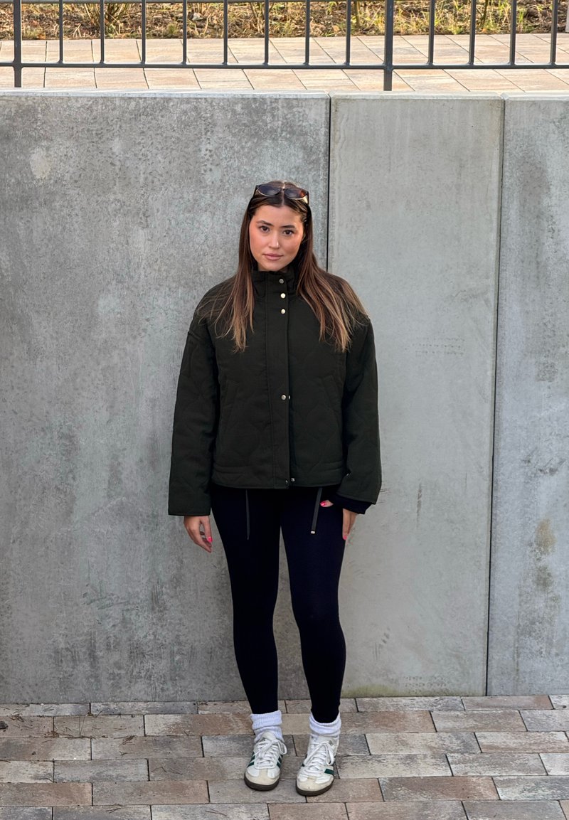 Veste matelassée verte avec col montant, accents de boutons dorés, assortie à des leggings noirs et des baskets blanches. Cheveux détachés, debout contre un mur en béton.