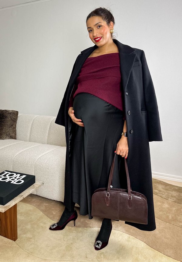 Tenue de maternité comprenant un pull bordeaux, une jupe noire, un manteau noir, un sac à main sombre et des escarpins rouges ornés à l'avant. Fond neutre.