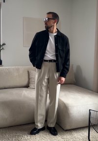 Άνδρας με μαύρο σακάκι, λευκό πουκάμισο, μπεζ παντελόνι και μαύρα παπούτσια, στέκεται σε εσωτερικό χώρο κοντά σε μπεζ καναπέ και λιτή τοιχογραφία.