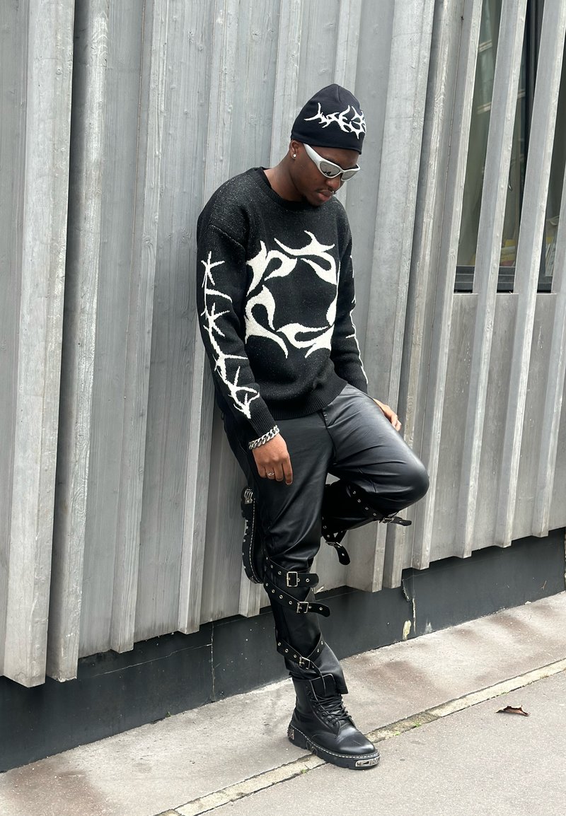 Black jumper with white abstract designs, black leather trousers with buckles, and black combat boots, accessorised with a black beanie and sunglasses.