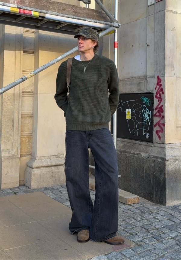 Uomo con maglione verde, berretto mimetico, jeans neri larghi e scarpe marroni sta in piedi con le mani dietro la schiena vicino a ponteggi e a un muro coperto di graffiti.