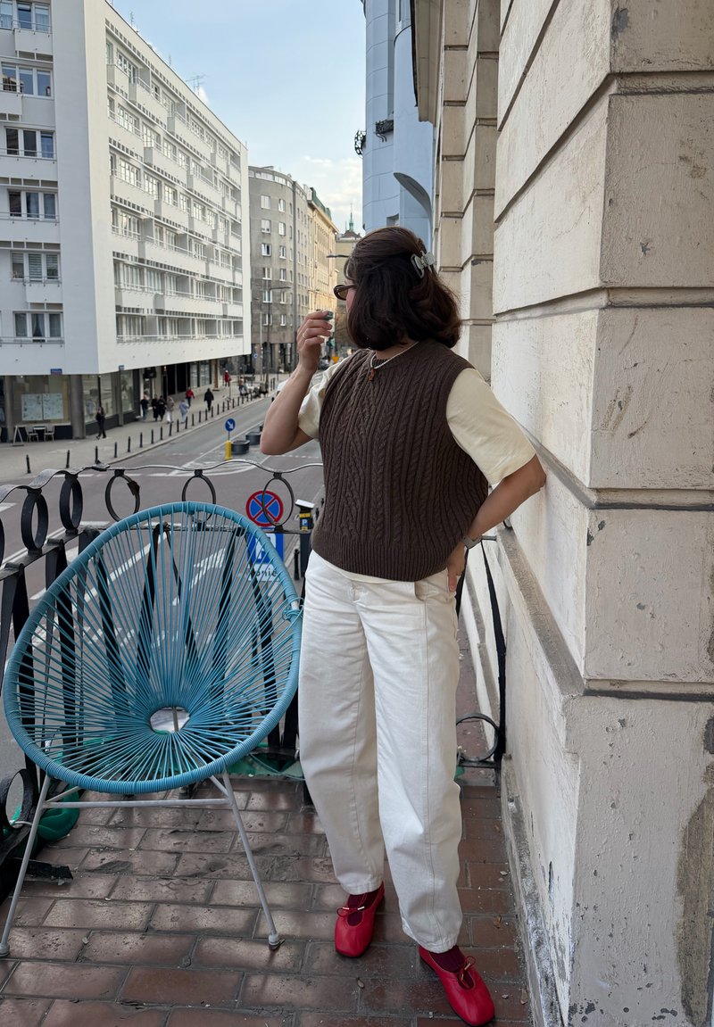 Person in brauner Strickweste, cremefarbenem Hemd, weißer Hose und roten Schuhen steht auf dem Balkon neben einem blauen Stuhl und blickt auf die Straße mit Gebäuden.