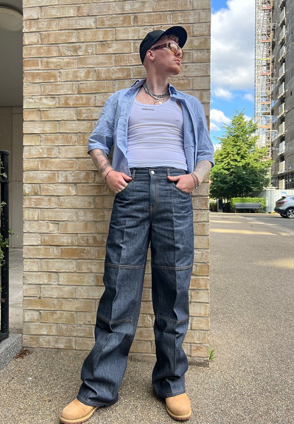 Pantalones de mezclilla de corte holgado, lavado oscuro y costuras en contraste. Combinados con una camisa de color azul claro y botas de color tostado.