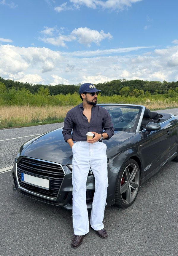 Hombre con gafas de sol y gorra apoyado en un coche descapotable negro al borde de la carretera sosteniendo una taza de café bajo un cielo parcialmente nublado.