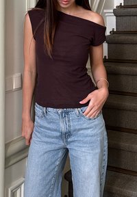 Sötétbarna, vállnélküli, testhez simuló felső, világoskék, magas derekú farmernadrággal párosítva. Sima anyag, laza dizájnnal. Egyszerű karkötő kiegészítő.