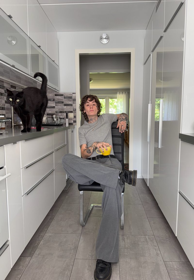 Personne avec des tatouages assise en tailleur sur une chaise dans une cuisine moderne tenant une tasse jaune, tandis qu'un chat noir marche sur le plan de travail.