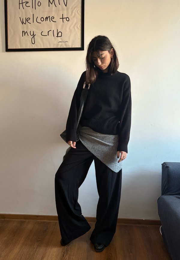 Maglione nero oversize, avvolgente asimmetrico grigio, pantaloni neri larghi, scarpe nere. Stanza con pavimento in legno e opere d'arte appese alle pareti sullo sfondo.