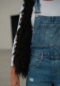 Mono de mezclilla con un bolsillo frontal, con un chal negro texturizado suelto sobre una camiseta blanca de manga larga. Los detalles incluyen herrajes plateados.