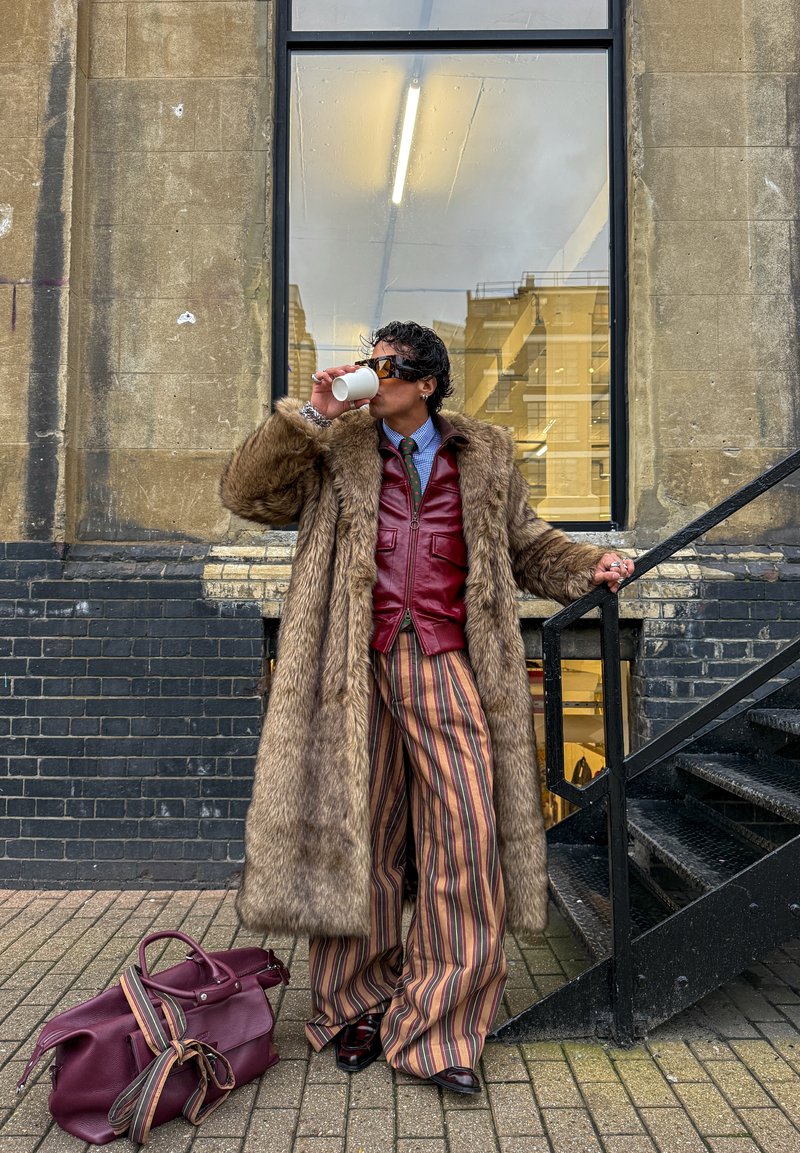 Personne portant un long manteau en fourrure marron, une veste en cuir rouge, un pantalon large rayé, des lunettes de soleil, buvant dans une tasse, debout près d’un escalier en métal noir.