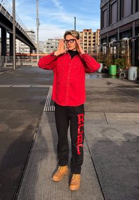 Jeune personne portant un t-shirt rouge et un pantalon noir avec des lettres rouges, debout sur un trottoir urbain, ajustant de grandes lunettes noires, journée ensoleillée avec des bâtiments en arrière-plan.