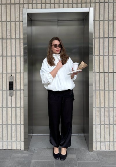 Mujer con chaqueta blanca y pantalones negros sosteniendo café y comida de pie en un ascensor con fondo de pared con azulejos.
