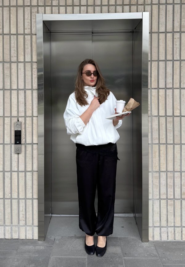 Frau in weißer Jacke und schwarzer Hose hält Kaffee und Essen und steht im Aufzug mit gefliestem Wandhintergrund.