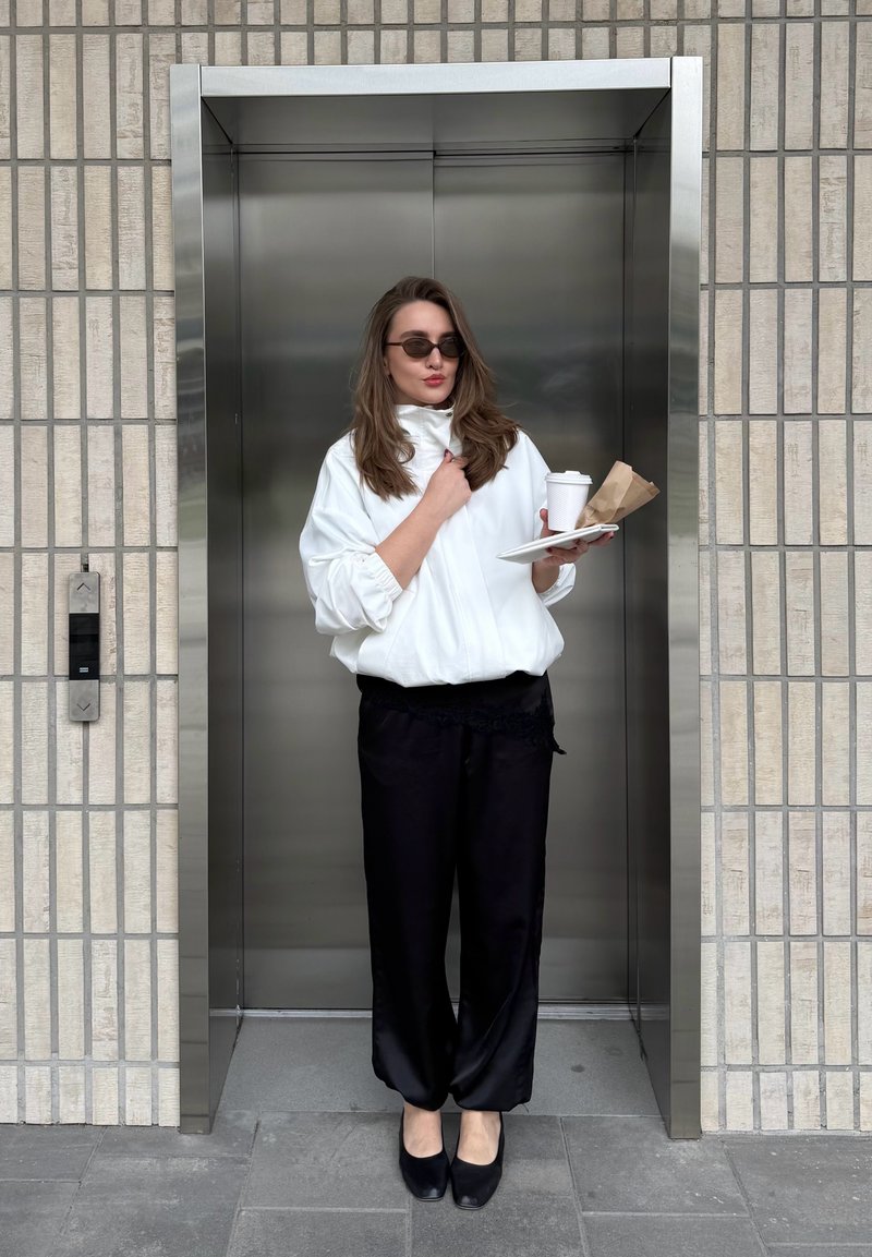 Frau in weißer Jacke und schwarzer Hose hält Kaffee und Essen und steht im Aufzug mit gefliestem Wandhintergrund.