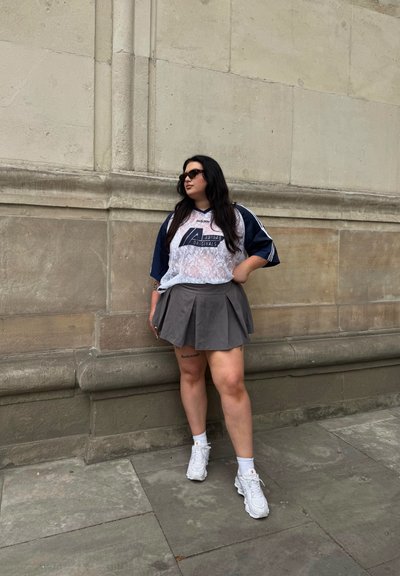 Camiseta blanca de encaje con detalles en azul marino, falda gris con pliegues y zapatillas blancas. La modelo lleva gafas de sol negras y está de pie frente a una pared de piedra.