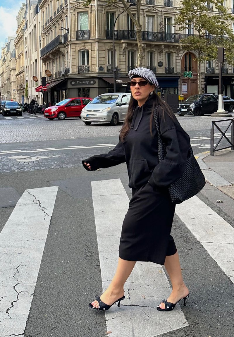 Hoodie noir surdimensionné, jupe midi noire, sandales à talons hauts à lanières, grand sac tissé, lunettes de soleil et casquette rayée noir et blanc.