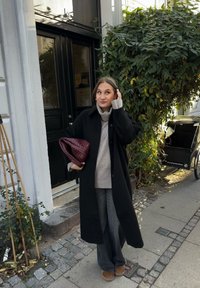 Čierny dlhý kabát cez sivý pletený sveter, široké sivé nohavice a hnedé čižmy. Drží bordovú tkáčsku tašku. V pozadí zelené listy.