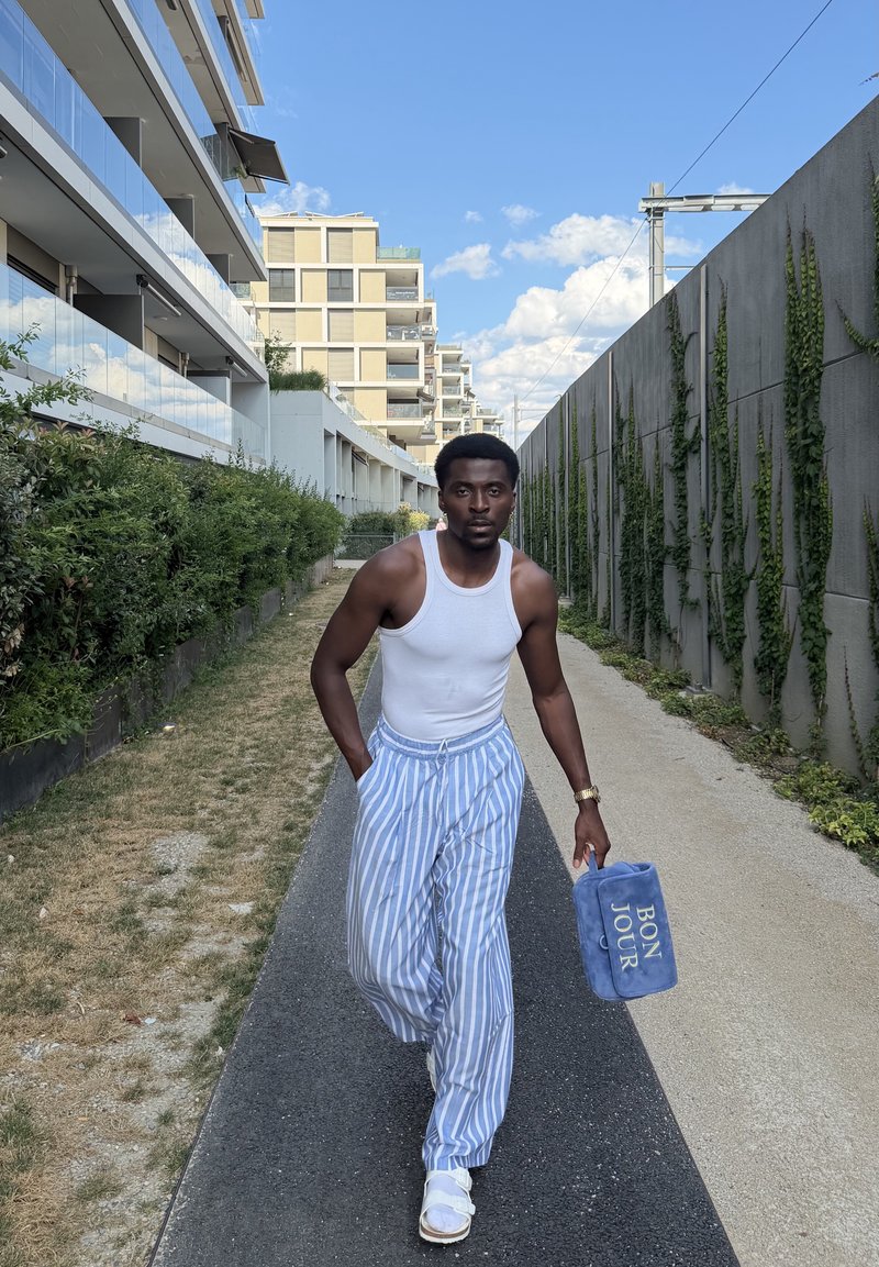 Homme marchant sur un chemin urbain entre des bâtiments, portant un pantalon à rayures, un débardeur blanc, des sandales, portant un sac bleu avec le texte "BON JOUR".