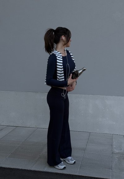 Mujer con cola de caballo, vistiendo pantalones y top negros, suéter de rayas sobre los hombros, sosteniendo una botella de agua, de pie sobre el pavimento frente a una pared gris.