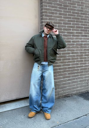 Hombre con vaqueros azules extragrandes, suéter marrón, chaqueta verde, gorra marrón y gafas, de pie contra una pared de ladrillos ajustándose las gafas.