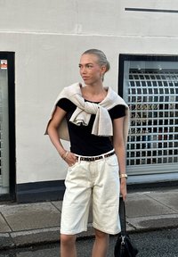 Μαύρο κοντομάνικο πουκάμισο, κρέμες πλεκτή ζακέτα ριγμένη στους ώμους, μπεζ σορτς με ζώνη και μαύρη τσάντα. Δρόμος με φόντο κτίριο.