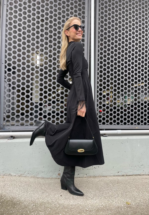 Black long-sleeve dress with a flowing silhouette and feathered cuffs. It is paired with knee-high black heeled boots and a structured black handbag.