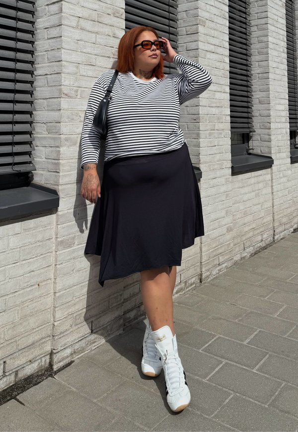 Femme aux cheveux roux portant un chemisier à manches longues rayé, une jupe noire, des baskets hautes blanches et des lunettes de soleil, appuyée contre un mur de briques blanches en plein air.