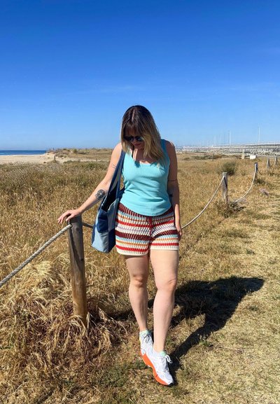 Mujer con gafas de sol, camiseta sin mangas azul, pantalones cortos a rayas y zapatillas blancas caminando por un camino de hierba seca cerca de la playa, llevando una bolsa azul.