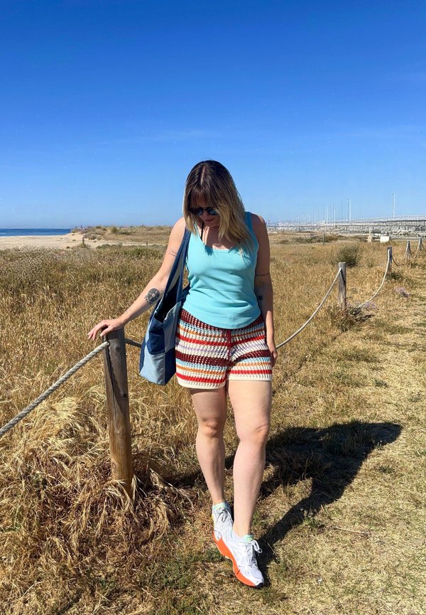 Kvinde med solbriller, blå tanktop, stribede shorts og hvide sneakers går på en tør grusstig ved stranden og holder en blå taske.