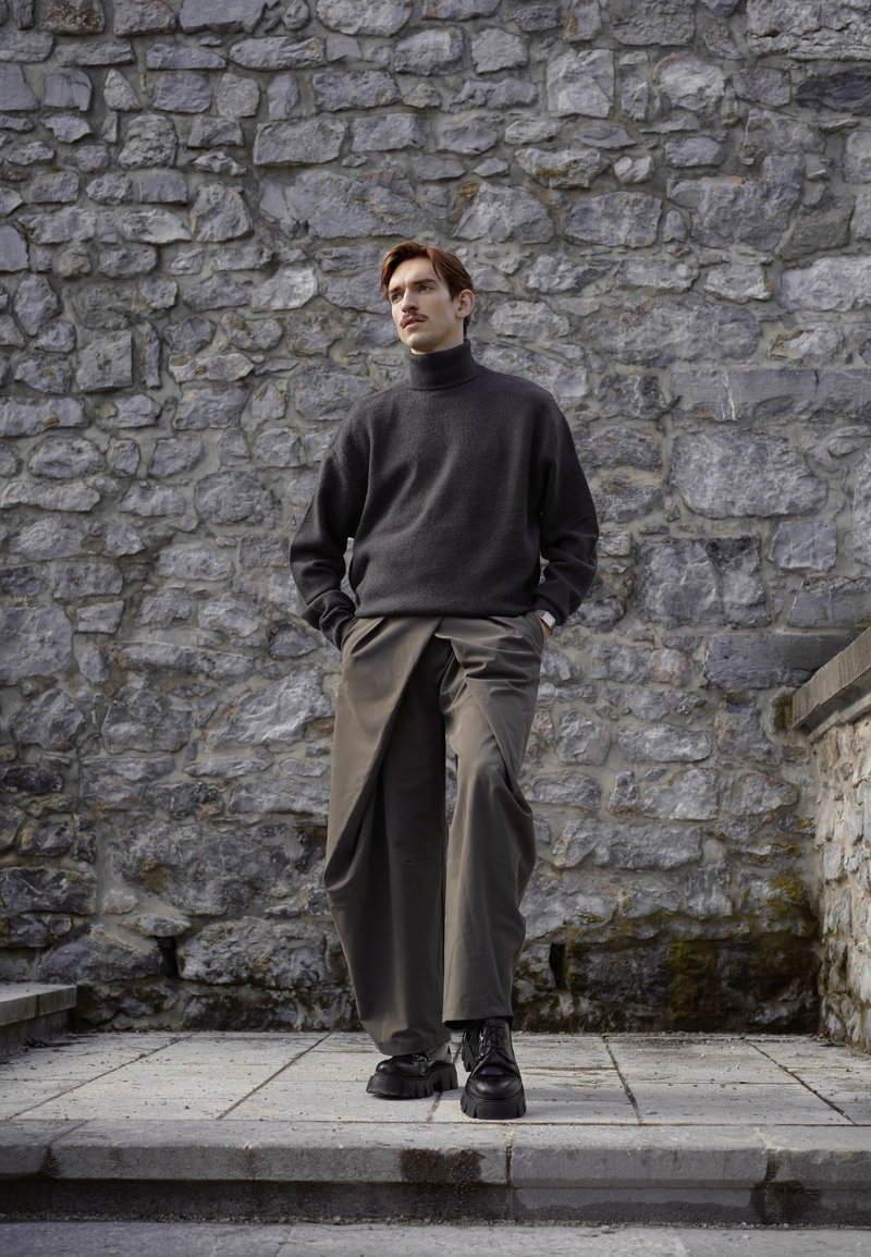 Jeune homme aux cheveux bruns portant un col roulé sombre, un large pantalon olive et des bottes noires, debout sur des marches en pierre devant un mur en pierre rugueux.