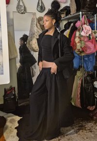 Woman with braided hair wearing loose black trousers, black top, and black coat in a room with racks of colourful coats and bags.