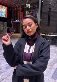 Musta takki päällä violetti ja valkoinen vetoketjullinen neulepaita. Henkilö pitelee viininpunaista silmälasiparia. Sisätila, jossa on moderni arkkitehtuuri ja tunnelmallinen valaistus.